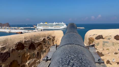 Eine-Kanone-über-Dem-Golf-Von-Oman-Bei-Muttrah-Fort-Mit-Einem-Kreuzfahrtschiff-Im-Hafen