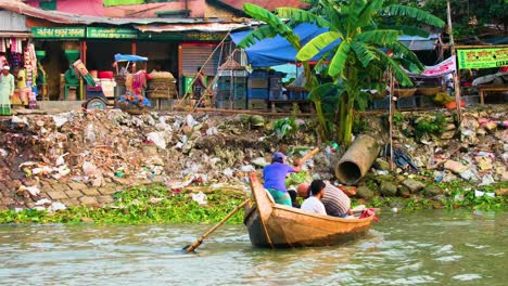 Boatman-rowing-on-polluted-river-near-littered-riverbank-with-onlookers-and-shops