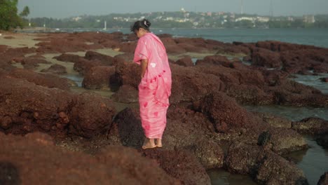 Woman-in-pink-saree-exploring-rocky-beach-shore,-city-in-distance,-sunny-day