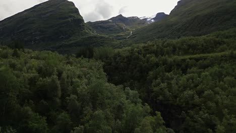 large-green-forest-in-a-valley-surrounded-by-mountains-with-a-waterfall-and-snow-on-the-peaks,-norway,-europe,-drone