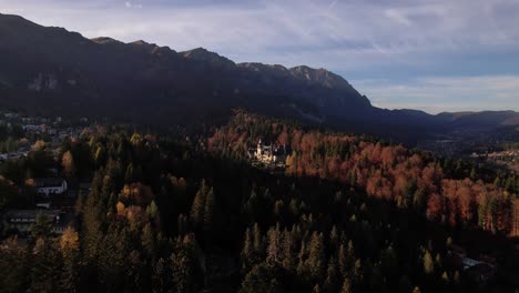 Peles-castle-amidst-autumn-colored-forest-with-bucegi-mountains-backdrop-at-sunset,-aerial-view