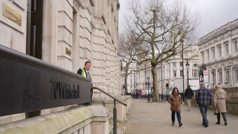 People-walk-along-Whitehall-as-a-security-guard-stands-outside-70-Whitehall,-the-entrance-to-the-UK-government-Cabinet-Office