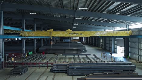 Aerial-tracking-pullback-follows-overhead-bridge-crane-moving-across-top-of-steel-beam-warehouse
