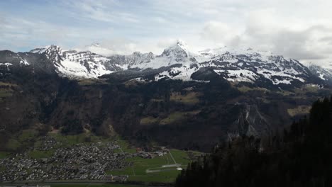 Panoramic-aerial-of-Glarus,-Switzerland,-a-quaint-residential-enclave-nestled-in-the-valley,-embraced-by-towering-snow-capped-peaks