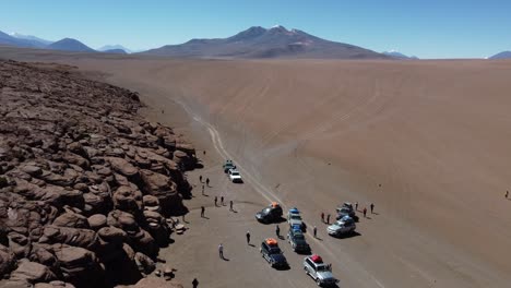 Aerial:-Tourism-vehicles-park-on-desert-sand-near-eroded-rock-bluff