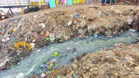 Water-stream-filled-with-trash-flowing-through-a-polluted-area,-people-and-buildings-in-the-background