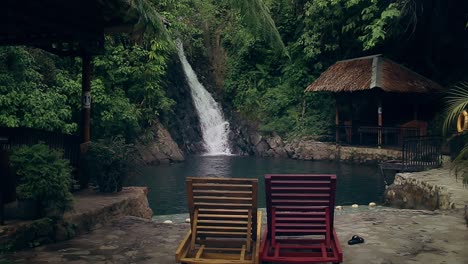 A-pair-of-two-wooden-lounge-chairs-facing-the-view-of-a-cascading-waterfall-with-a-native-hut-beside-it,-Pulangbato-Falls-in-Valencia,-Negros-Oriental-Philippines