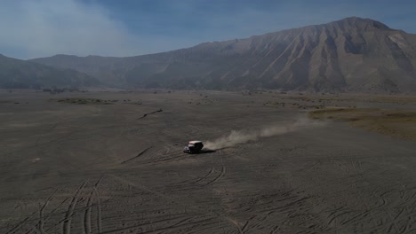 Following-a-car-with-drone-through-Bromo's-lava-fields-at-sunrise,-with-striking-sand-rush-tire-tracks