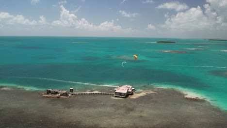 Kitesurfers-near-a-palafito-in-los-roques-during-a-sunny-day,-aerial-view