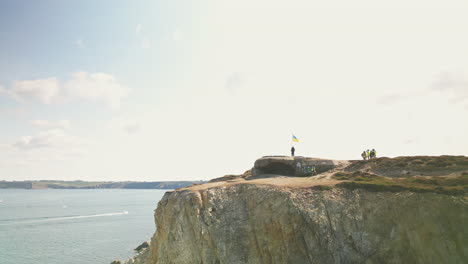 Enthüllende-Bogenantenne-Eines-Mannes,-Der-Auf-Einem-Verlassenen-Bunker-Auf-Einer-Klippe-Mit-Blick-Auf-Das-Wasser-Mit-Ukrainischer-Flagge-Steht