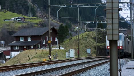 Tren-De-Carga-Acercándose-En-El-Paisaje-Montañoso-Suizo,-Casas-En-El-Fondo,-Durante-El-Día