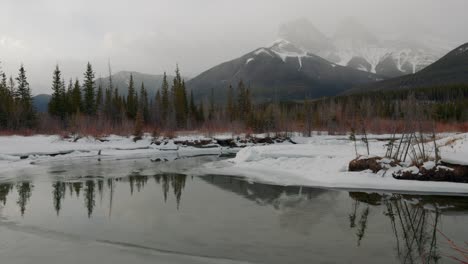 Foggy-morning-in-Canmore,-Alberta,-Canada-with-the-iconic-Three-Sisters-mountains-shrouded-in-snow,-while-a-tranquil-pond-graces-the-foreground,-offering-a-glimpse-into-the-ethereal-beauty-of-nature