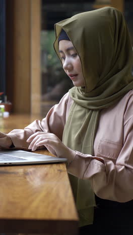 Jóvenes-Hermosas-Mujeres-Musulmanas-Asiáticas-Modernas-Sentadas-Junto-A-La-Ventana-Disfrutando-De-Un-Momento-Relajante-Trabajando-En-Una-Computadora-Portátil-En-La-Cafetería-En-Un-Día-Soleado