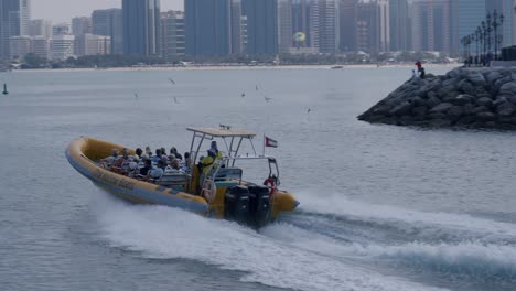 Turistas-En-Lancha-Rápida-Navegando-Frente-Al-Horizonte-Del-Paisaje-Urbano-De-Abu-Dhabi-Y-Los-Edificios-Frente-Al-Mar,-La-Compañía-De-Cruceros-Yellow-Boats