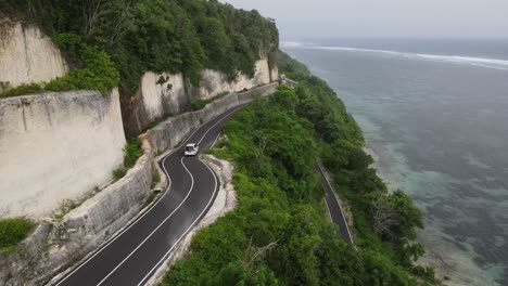 Aerial-view,-a-white-car-driving-along-the-cliff-side-asphalt-road-and-the-sea-located-on-Tanah-Barak-beach,-Bali-island,-Indonesia