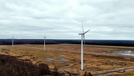 group-of-tall-white-wind-turbines-with-three-long-blades-standing-in-a-vast-green-field-under-a-cloudy-sky,-slowmo,-Aerial-Pan-Shot