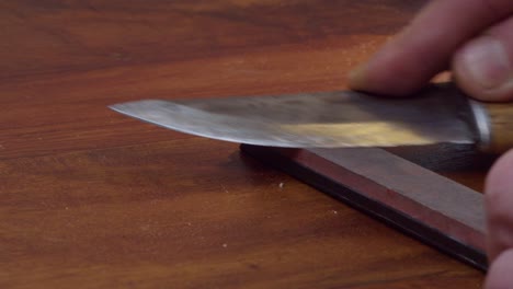 Man-sharpens-knife-edge-on-whetstone-on-wooden-counter,-tool-close-up
