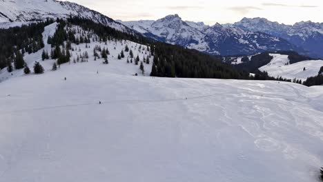 Vast-snowy-landscape-in-Switzerland-with-distant-skiers-and-mountain-backdrop,-aerial-view