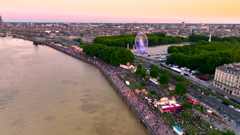 Feria-Del-Vino-En-La-Orilla-Del-Río-Garona-Con-Noria-Y-Multitudes-Al-Atardecer,-Inclinación-Aérea-Hacia-Abajo-Y-Tiro-Ascendente