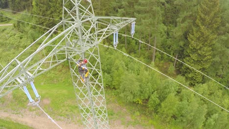 Worker-climbing-high-voltage-tower-in-forest,-aerial-view