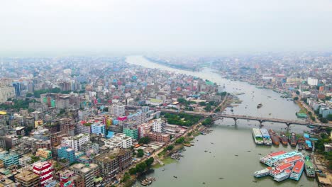 Descending-drone-shot-of-Dhaka,-Bangladesh-with-buildings-and-river-with-boat-in-buriganga