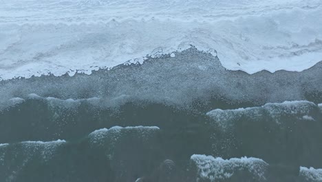Foamy-waves-lapping-at-the-frozen-edge-of-a-serene-lake,-captured-in-the-dim-light-of-dusk