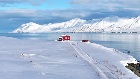 Beautiful-scenery-of-a-single-red-house-on-the-edge-of-a-fjord,-surrounded-by-snow-and-mountains