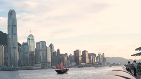 Blick-Auf-Das-Ikonische-Hölzerne-Dschunkenboot-Mit-Roten-Segeln,-Bekannt-Als-Aqua-Luna,-Das-über-Den-Victoria-Harbour-Und-Die-Skyline-Von-Hongkong-Segelt,-Während-Der-Sonnenuntergang-Beginnt