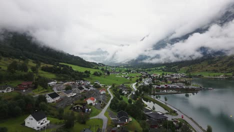 Pequeño-Pueblo-En-Un-Lago-O-Fiordo,-Valle-Verde-Con-Altas-Montañas-Y-Nubes-Al-Fondo,-Noruega,-Naturaleza,-Drone