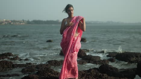 Woman-in-pink-sari-stands-on-rocky-shore,-shielding-eyes-to-gaze-out-to-sea,-serene-ocean-backdrop