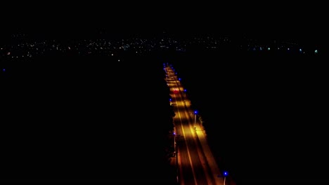 Vista-Nocturna-De-Una-Carretera-Iluminada-Con-Autos,-Que-Se-Extiende-Hacia-Un-Paisaje-Urbano-Bajo-Un-Cielo-Oscuro,-Vista-Aérea