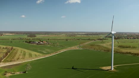A-single-wind-turbine-in-green-rural-landscape-on-a-clear-day,-long-shadows-cast-on-the-ground,-aerial-view