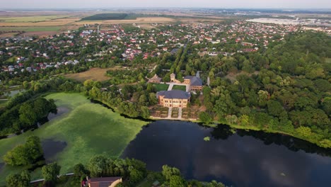 Palacio-Mogoșoaia-Con-Lago-Adyacente-Y-Vegetación-Durante-La-Luz-De-La-Mañana,-Las-Afueras-De-La-Ciudad-Al-Fondo,-Vista-Aérea