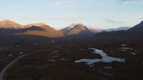 Panoramic-drone-landscape-mountains-at-Isle-of-Skye,-Scotland-travel-destination-wet-agricultural-fields-and-road-through-natural-environment