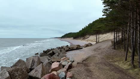 Rocky-ocean-beach-with-rough-grey-triangle-cliffs-on-either-side-Green-trees-growing-on-Baltic-beach-Aerial-Left-Truck-Shot