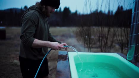 Man-With-Water-Hose-Filling-Up-The-Hot-Tub-Over-Wood-Fire-Outside-His-Cabin