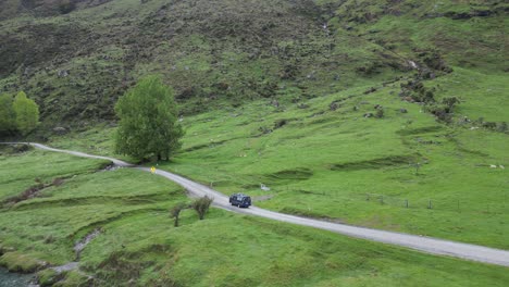 A-van-drives-along-a-gravel-road,-its-journey-captured-by-a-drone,-revealing-scenic-landscapes-of-trees,-streams,-and-grazing-sheep-in-the-remote-countryside