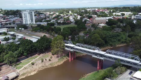 Aerial-view-of-a-pedestrian-bridge-over-a-brown-river-approaching-a-green-field-and-a-large-Catholic-Church-in-Australia