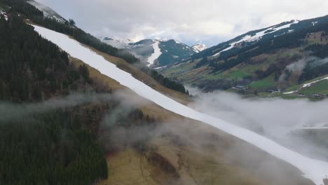 Nebel-über-Dem-Skigebiet-Saalbach-Hinterglemm,-Schneebedeckte-Berge-Flankieren-Ein-Grünes-Tal,-Luftaufnahme