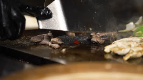 Spatula-flips-sizzling-beef-fajita-strips-on-flat-top-grill,-chicken-and-beef-fajitas-with-peppers-and-onions-cooking-in-commercial-restaurant-kitchen-griddle,-slow-motion-close-up-4K