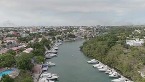 Aerial-forward-over-canal-of-La-Romana-port-with-boats-anchored-along-river-shores,-Dominican-Republic