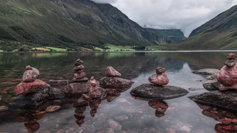 Peculiares-Mojones-De-Piedra-Se-Encuentran-En-El-Borde-Del-Lago-Poco-Profundo,-Rodeados-De-Montañas-Cubiertas-De-Bosque