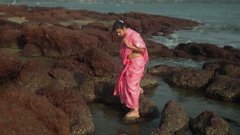 Young-girl-in-pink-dress-exploring-rocky-shoreline,-waves-crashing-nearby,-daylight-adventure