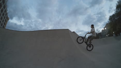 BMX-biker-huge-air-out-of-skatepark-bowl-quarter-pipe-hip,-bike-trick-flying-in-the-air-at-dusk-in-extreme-800fps-slow-motion,-4k-fisheye