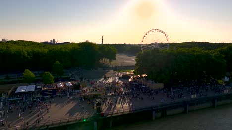 Feria-Del-Vino-Con-Noria-Y-Multitudes-Mientras-El-Sol-Se-Pone-Detrás,-Toma-Aérea-A-La-Izquierda