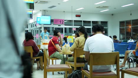 Passagiere-Sitzen-Vor-Einem-Fernsehbildschirm,-Auf-Dem-Die-Abfahrtszeiten-Der-Busse-Zu-Verschiedenen-Zielen-In-Thailand-Angezeigt-Werden