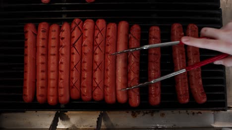 Hand-With-Tongs-Cooking-And-Rolling-Hot-Dogs-On-Grill
