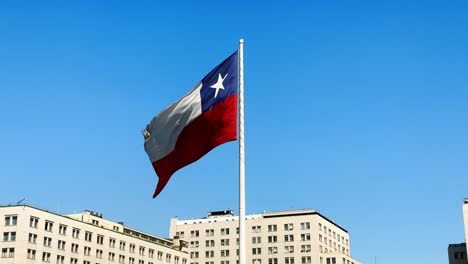 Bandera-Chilena-Ondeando-Orgullosamente-Contra-Un-Cielo-Azul-Claro,-Sobre-El-Telón-De-Fondo-Del-Paisaje-Urbano