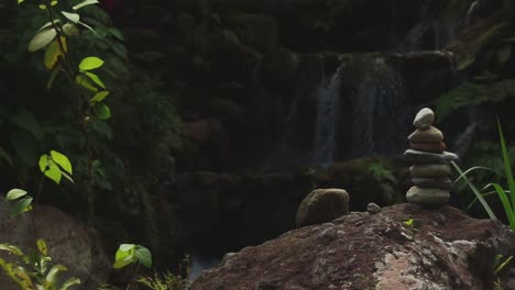 A-serene-scene-with-a-stack-of-stones-in-front-of-a-gentle-waterfall-surrounded-by-lush-greenery