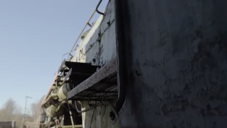 close-up-of-the-weathered-side-of-an-old-green-train-car-with-rivets-and-peeling-paint,-next-to-a-rusty-metal-wheel-on-railway-tracks,-in-a-sunny-train-yard-with-a-clear-blue-sky-in-the-background